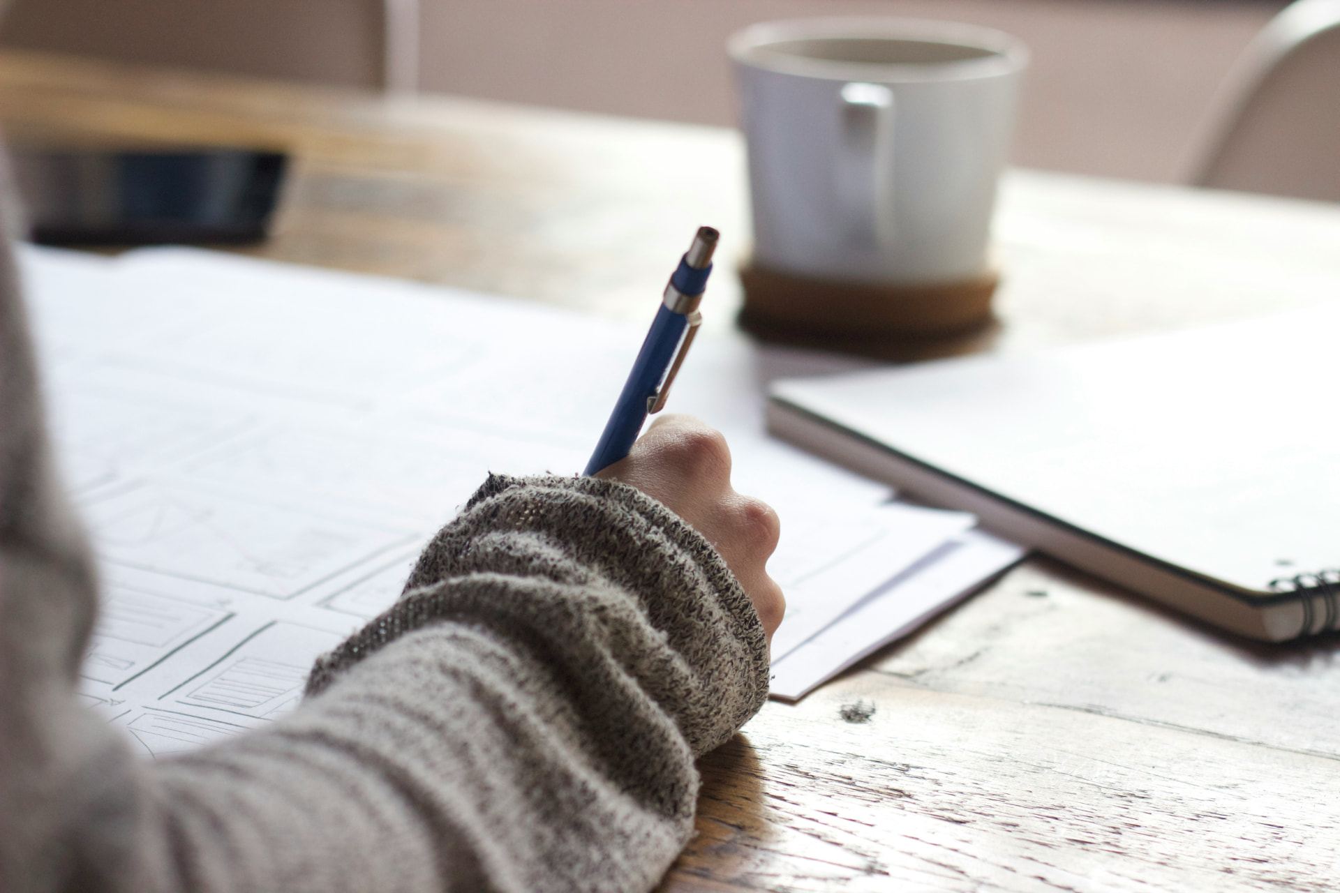 A hand sketching a plan in a notebook beside a cup of coffee